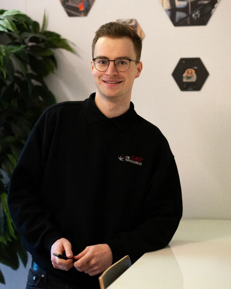 Mitarbeiter der Eventhandwerker mit Brille und schwarzem Firmenpullover steht an einem Empfangstresen.