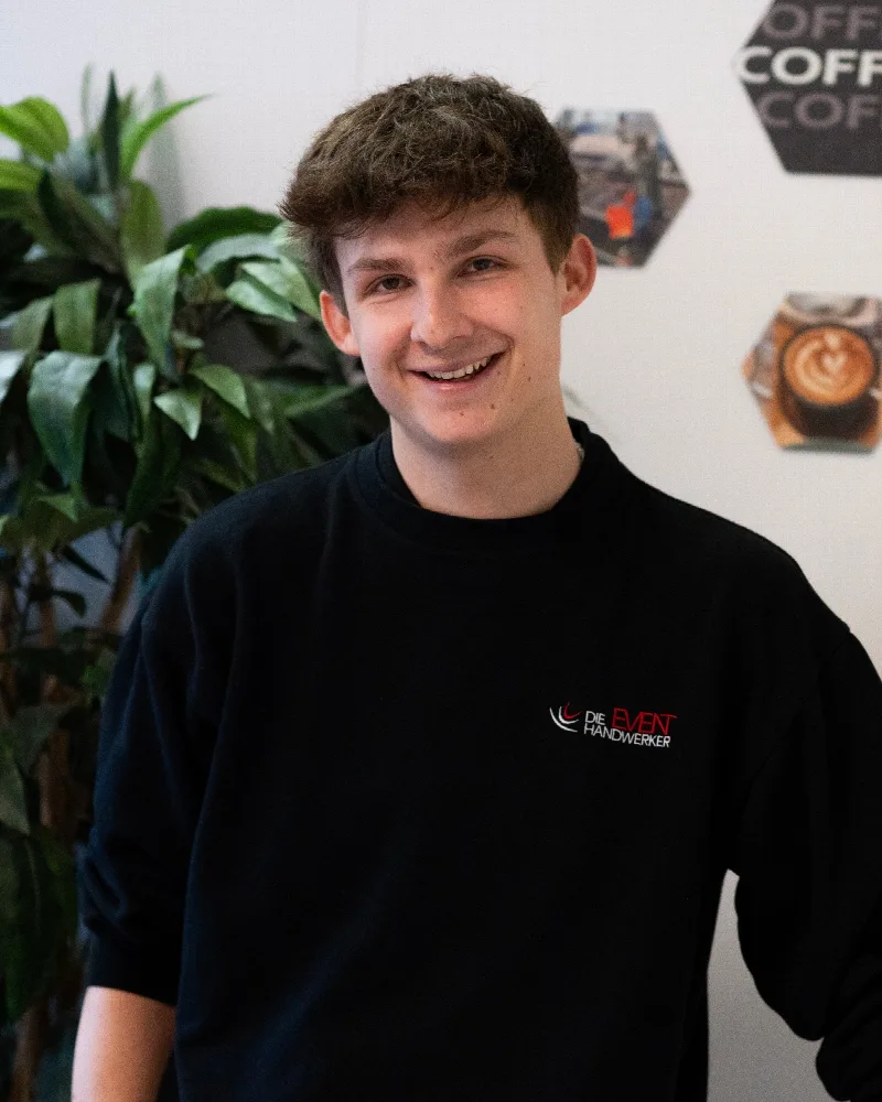 Lächelnder Mitarbeiter der Eventhandwerker in schwarzem Pullover vor einer Pflanze und einer Wand mit Kaffeebildern.