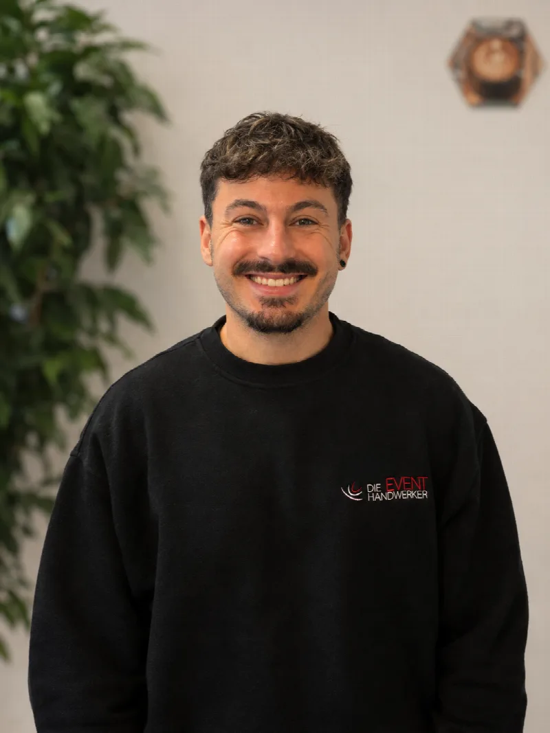 Lächelnder Mitarbeiter der Eventhandwerker in einem schwarzen Sweatshirt vor einer hellen Wand.